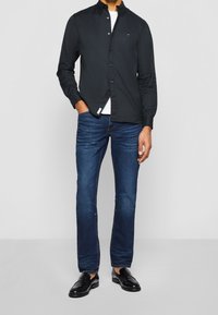 Musta kauluspaita, valkoinen T-paita alla, siniset farkut, mustat nahkakengät. Paidassa on pieni logo rinnassa.