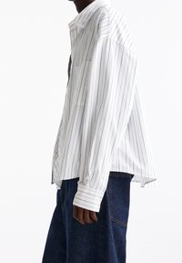 Personne portant une chemise blanche ample à fines rayures avec boutons et un jean en denim bleu foncé, debout de profil sur un fond blanc uni.