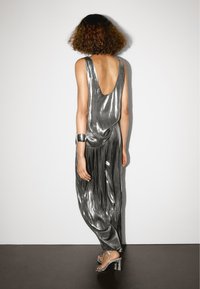 Metalický stříbrný overal s nízkým zadním dílem a splývavým designem. Má volný střih, hladkou texturu a je kombinován s sandály na vysokém podpatku se zavazováním.