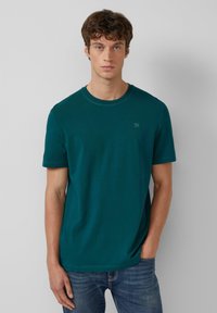 Teal katoenen T-shirt met een ronde hals, korte mouwen en een klein logo op de borst, gecombineerd met blauwe spijkerbroeken. Soepele textuur.