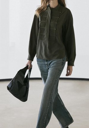Femme marchant à l'intérieur portant un chemisier à manches longues sombre avec des plis et des perles, un jean bleu clair, portant un sac à main patchwork noir.