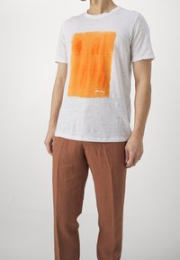 Weißes kurzärmeliges T-Shirt mit einem orangefarbenen abstrakten Design. Kombiniert mit locker sitzenden, braunen Leinenhosen. Einfacher, minimalistischer Stil.