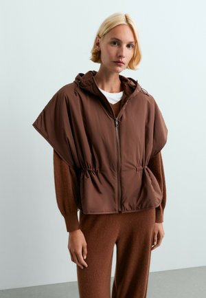 Femme blonde portant un gilet marron à manches larges zippé par-dessus un pull et un pantalon marron assortis, debout devant un fond clair uni.