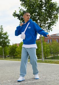 Blå fleece hettegenser med lommer foran, lagd over en hvit t-skjorte, kombinert med lyseblå løse jeans og hvite sneakers med blå detaljer.
