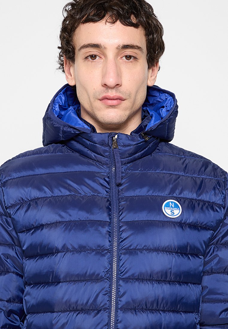 Giaccone imbottito blu navy con una texture lucida, dotato di zip frontale, cappuccio e un logo circolare blu sul lato sinistro del petto. Design a righe orizzontali.