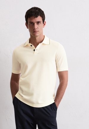 Lichtgele polo met korte mouwen, voorzien van drie zwarte knopen, een contrasterende kraag en een gestructureerde stof. Draagt met donkere broek.