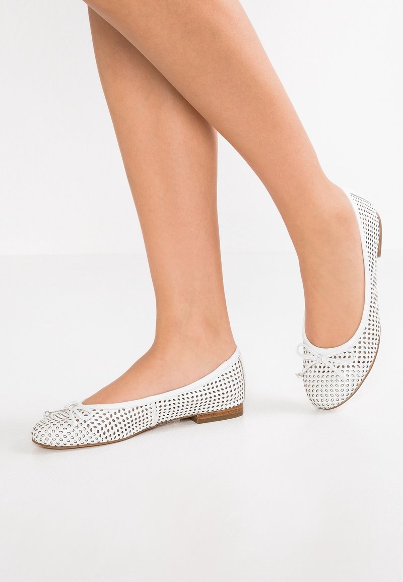 Jambes portant des ballerines blanches avec des découpes circulaires et de petits nœuds sur un fond blanc uni.