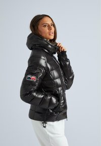 Frau in einer glänzenden schwarzen Steppjacke mit Kapuze, Hand in der Tasche, und einem bunten Patch auf dem linken Ärmel, leicht nach rechts gewandt.