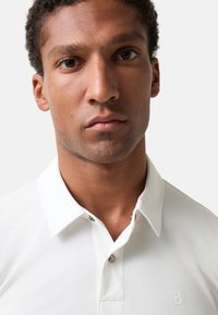 Nahaufnahme eines Mannes mit kurzen lockigen Haaren, der ein weißes Hemd mit Kragen und einem kleinen "B"-Logo auf der Brust trägt, vor einem einfarbigen Hintergrund.