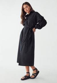 Schwarzes Hemdkleid mit langen Ärmeln, tailliertem Bund und seitlichen Schlitzen. Glattes Material mit matter Oberfläche. Kombiniert mit schwarzen, Riemchen-Sandalen.