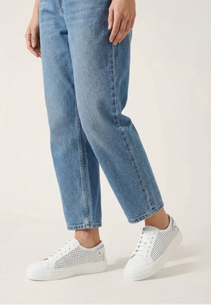 Person trägt hellblaue Jeans mit geradem Bein und weiße Sneakers mit perforierten Seitenpanelen, steht vor hellem Hintergrund.