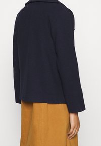 Veste en laine mélangée bleu marine avec un col structuré, un design cropped et des manches larges, associée à une jupe beige taille haute.