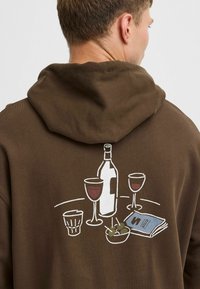 Brauner Kapuzenpullover mit einem grafischen Design eines Weins, Gläsern und einem Teller mit Oliven, alle in Weiß umrandet. Weicher Stoff.