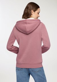 Felpa con cappuccio rosa caratterizzata da un interno morbido e foderato in peluche e polsini a costine. Indossata con jeans blu, che mettono in risalto un design casual.