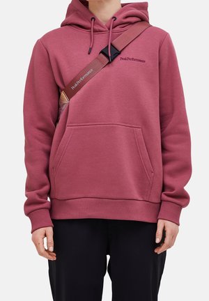 Personne portant un sweat à capuche rose foncé avec une poche avant et une bandoulière de sac marron portant l'étiquette Peak Performance.