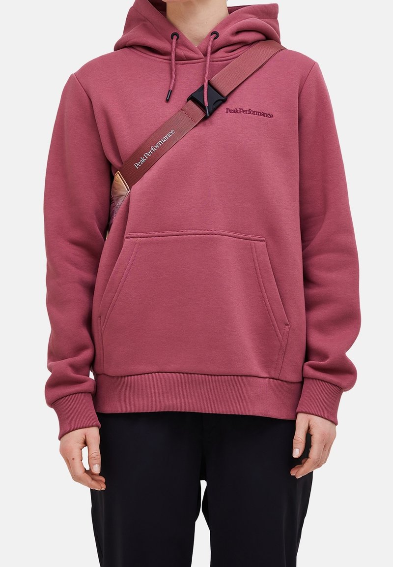 Personne portant un sweat à capuche rose foncé avec une poche avant et une bandoulière de sac marron portant l'étiquette Peak Performance.