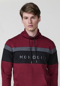Červená a čierna mikina s vysokým golierom, s vodorovnými pruhmi a prednou kapsou. Logo "KORÖSHI" v šedej farbe, šnúrky na úpravu.