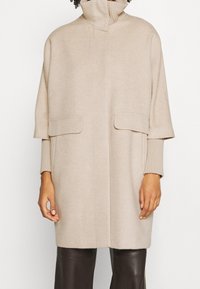 Beige wollen jas met een hoge kraag, korte mouwen en twee voorkeurszakken. Gladde textuur met een licht oversized pasvorm.