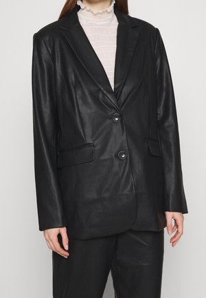 Kobieta ubrana w czarną skórzaną marynarkę na dwa guziki z przednimi kieszeniami z klapkami, na białą bluzkę z wysokim kołnierzem i falbanami.