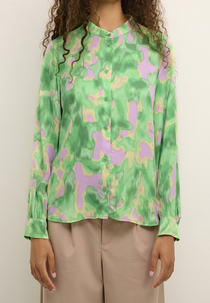 Mujer vistiendo una blusa de botones de manga larga con estampado abstracto en verde, púrpura y beige, combinada con pantalones anchos beige.