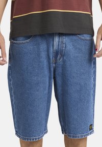 Mann trägt blaue Jeansshorts und ein zweifarbiges braun-schwarzes Hemd, zeigt die rechts rot lackierten Fingernägel.