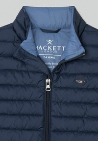 Granatowa pikowana kamizelka z wysokim kołnierzem i srebrnym zamkiem błyskawicznym. Na lewej piersi znajduje się czarne logo w kształcie diamentu.