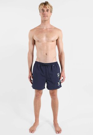 Homme blond debout pieds nus, torse nu, portant un short de bain bleu marine avec des imprimés floraux bleu clair et beige sur le côté gauche, sur fond blanc.