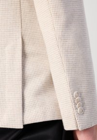 Giacca beige con texture a motivo houndstooth, dotata di un spacco e quattro bottoni chiari sui polsini. Vestibilità sartoriale.