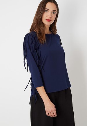 Top azul marino con mangas tres cuartos, que presenta acentos de flecos texturizados a lo largo de los hombros. Fabricado con un material suave y elástico.