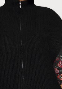 Haut noir en maille avec un col montant et une fermeture éclair centrale. Le tissu présente une texture côtelée, avec des manches à motif floral visibles.