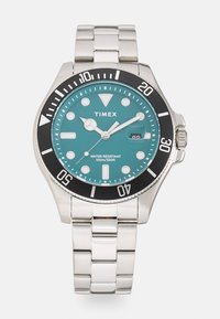 Reloj Timex con esfera turquesa, brazalete de acero inoxidable, bisel negro y marcas horarias blancas. Resistente al agua hasta 100m.