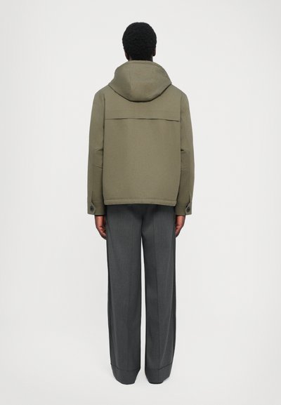 Olivgrüne Kapuzenjacke mit geradem Schnitt, mit einer Rückentasche, kombiniert mit grauen weit geschnittenen Hosen. Glatte Textur, minimalistisches Design.