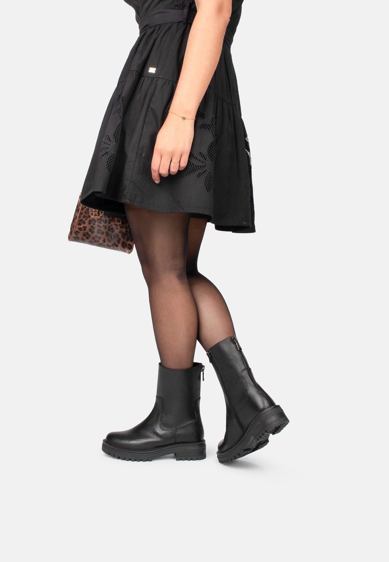Femme portant des bottines noires, des collants noirs transparents, une jupe noire avec des détails brodés, et tenant un sac à main imprimé léopard.