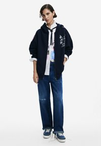 Marineblaue Kapuzenjacke mit Frontreißverschluss, seitlichen Taschen und Grafikdruck; darüber getragen mit einem weißen Shirt. Kombiniert mit ausgewaschenen blauen Jeans und Sneakers.