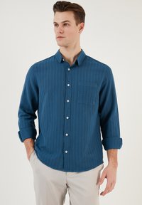 Cămașă button-up cu dungi albastre, guler clasic, mâneci lungi rulate la manșete și un buzunar pe piept. Textură moale a materialului și un luciu subtil.