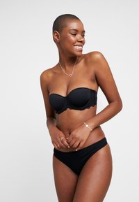 Zwarte strapless beha met kantafwerking, gladde cups en bijpassende zwarte bikini slip. Model poseert lachend en toont het ontwerp en de pasvorm.