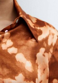 Camicia a maniche lunghe con colletto in tie-dye marrone e crema, con bottoni bianchi, indossata da una persona vista dal collo a metà petto su uno sfondo neutro.
