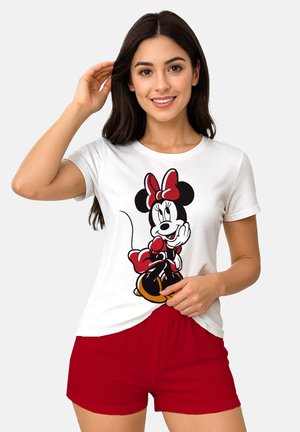 Giovane donna con lunghi capelli scuri che indossa una maglietta bianca con una stampa di Minnie Mouse e pantaloncini rossi, sorridente davanti a uno sfondo semplice.