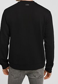 Hombre vistiendo una sudadera negra de Karl Lagerfeld con el logo en la parte superior de la espalda, combinada con jeans gris desgastados, visto desde atrás.