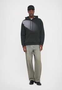 Mladostnik v črni kapi, črni hoodie, sivih ohlapnih kavbojkah in črnih čevljih, ki stoji ob enobarvni beli podlagi.