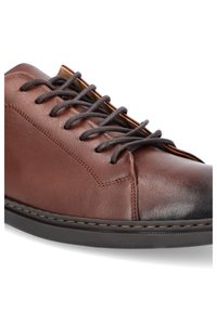 Brązowy skórzany but z ciemnymi sznurówkami, o niskim profilu, gładkich i teksturowanych powierzchniach, z kontrastującym noskiem oraz pikowanymi detalami.