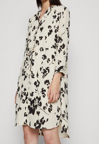 Nainen kermanvärinen nappipuseromekko, jossa on musta kukkakuosi. Mekossa on pitkät hihat, sidottava vyötärö ja hieman kaareva helma. Silkkinen kangas.