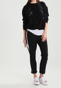 Zwarte geribbelde trui met een gestructureerd bloemenmotief, gecombineerd met zwarte tapered broek met witte zijkanten en witte sneakers.