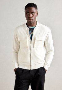 Chaqueta bomber blanca texturizada con cierre de cremallera y dos bolsillos en el pecho, combinada con una camisa de estampado colorido y pantalones negros.