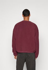 Sudadera de forro polar burdeos con superficie texturizada, hombros caídos y cuello redondo, combinada con jeans gris oscuro.