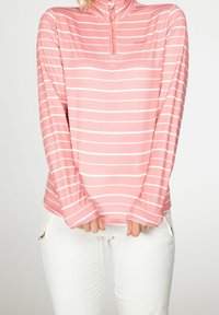 Mujer vistiendo una camiseta de manga larga a rayas horizontales en rosa y blanco con cremallera de un cuarto, combinada con pantalones blancos con bolsillos con cremallera.