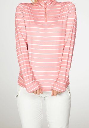 Donna che indossa un top a righe orizzontali rosa e bianche con maniche lunghe e zip quarti, abbinato a pantaloni bianchi con tasche a zip.