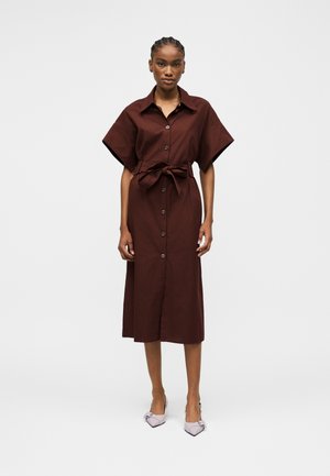 Vrouw staat in een bruine midi-jurk met knopen, een in de taille gestrikte riem en korte wijde mouwen, en draagt lichtgekleurde puntige hakken.
