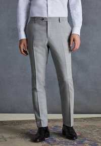 Graue maßgeschneiderte Hose mit feiner Textur, mit flacher Vorderseite, seitlichen Taschen und schwarzem Metallbeschlag, kombiniert mit schwarzen Schnürschuhen.