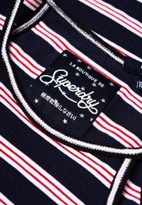 Marineblauwe stof met witte en rode horizontale strepen. Label met "Superdry" logo in witte schrift met accenten en sterren. Zwarte afwerking.